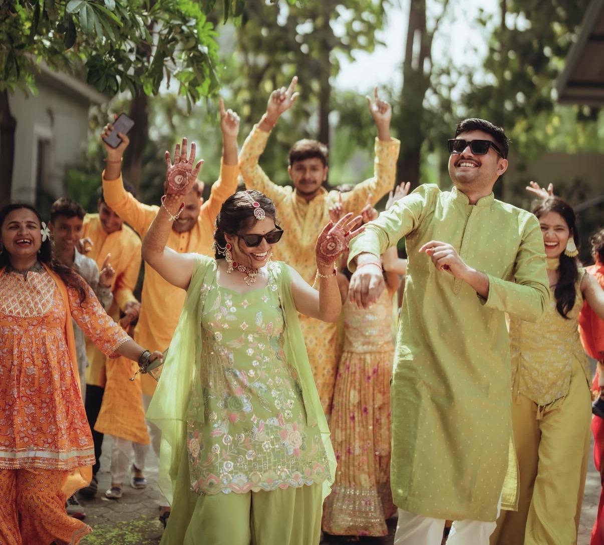 A couple in green dancing with their families in yellow at a daytime haldi