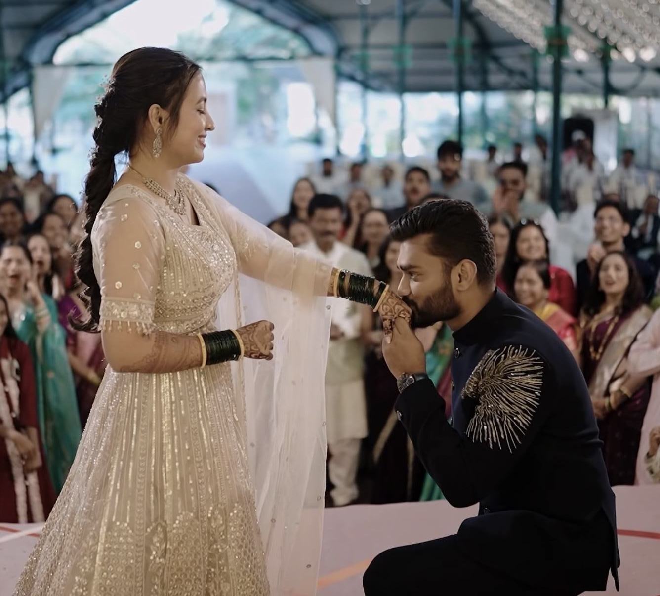 A groom on one knee kissing the bride's hand as the sangeet crowd watches