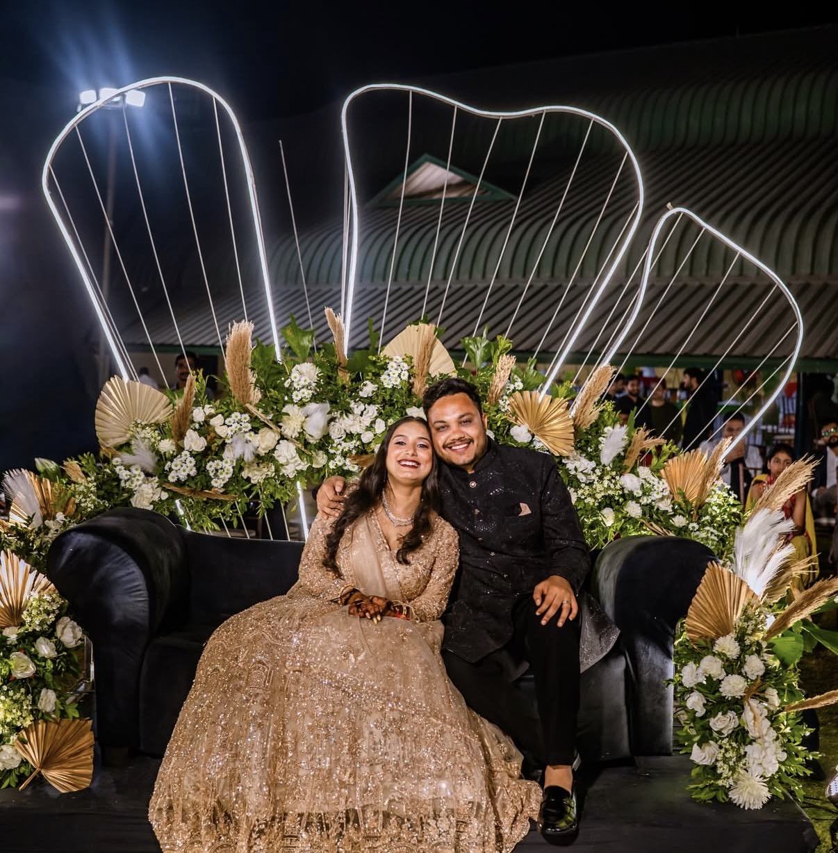 A couple on a black sofa in front of lit harp-shaped arches and white florals