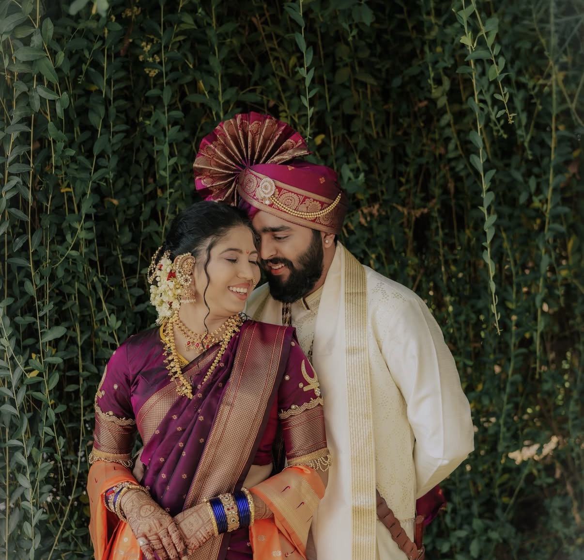 A bride in a purple Paithani and a groom in a pheta, laughing together against a wall of green