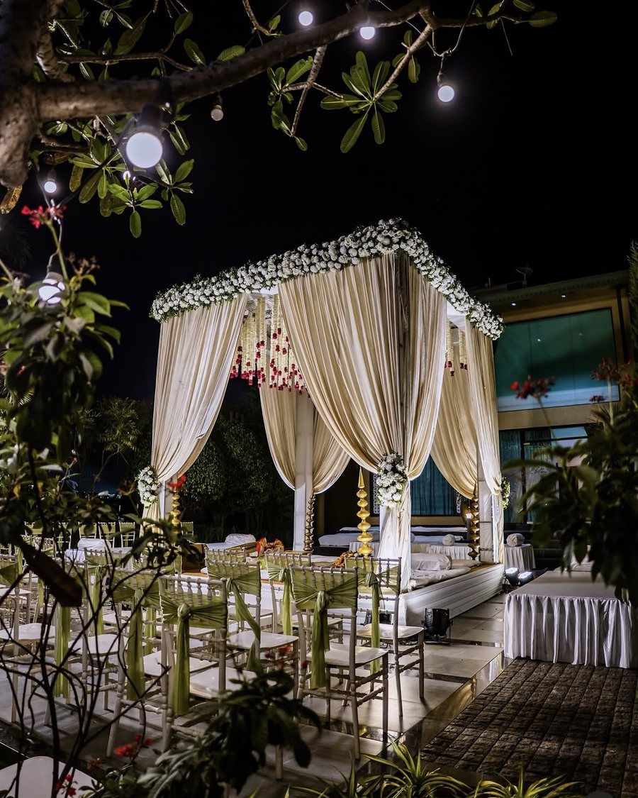 A night mandap of cream drapes and white florals under a frangipani tree strung with bulbs, the chairs sashed in green