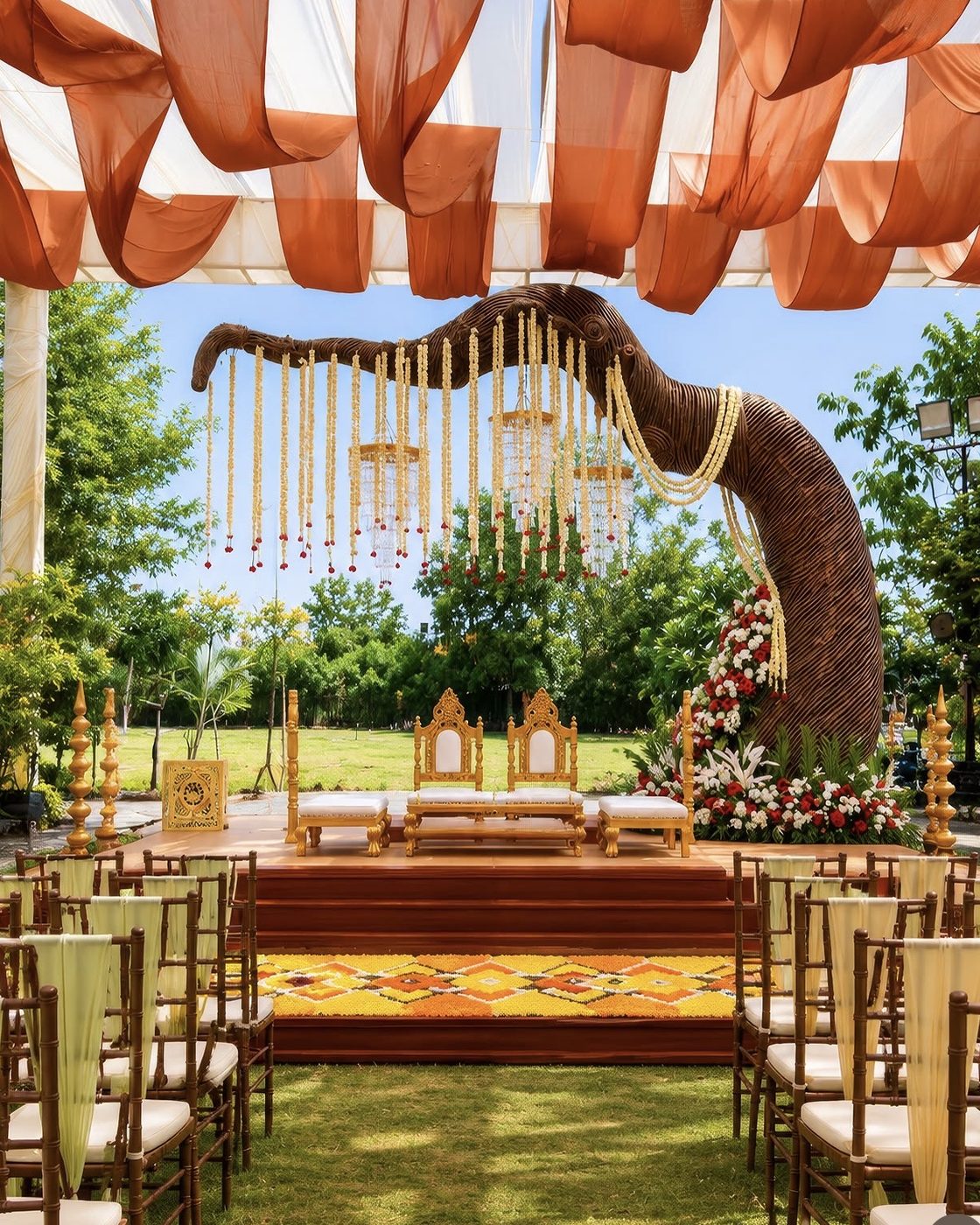 A daytime mandap under orange and white drapes, with a carved elephant-trunk arch and hanging floral strings