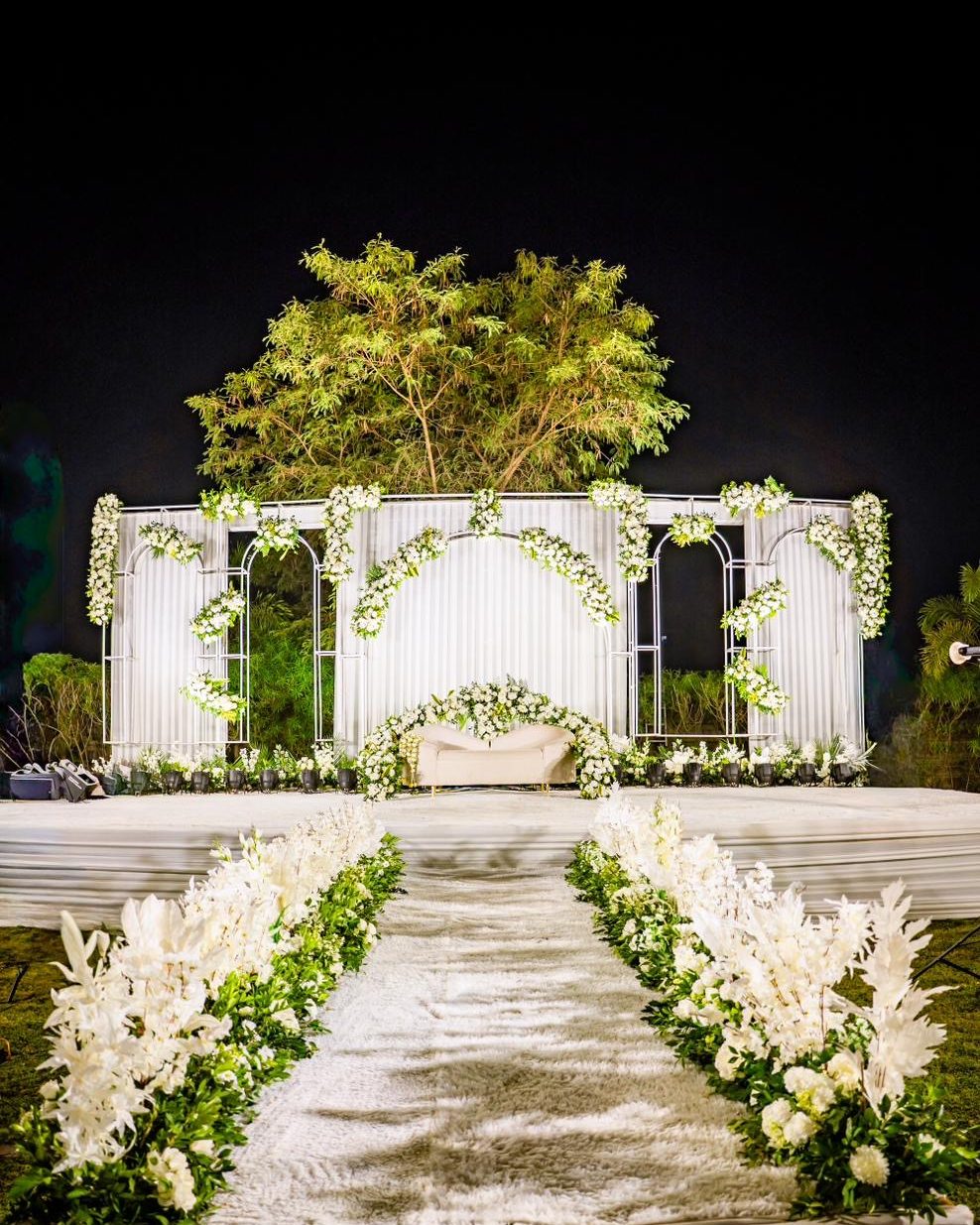 A night stage in white: lit arches, white florals and a flower-lined aisle