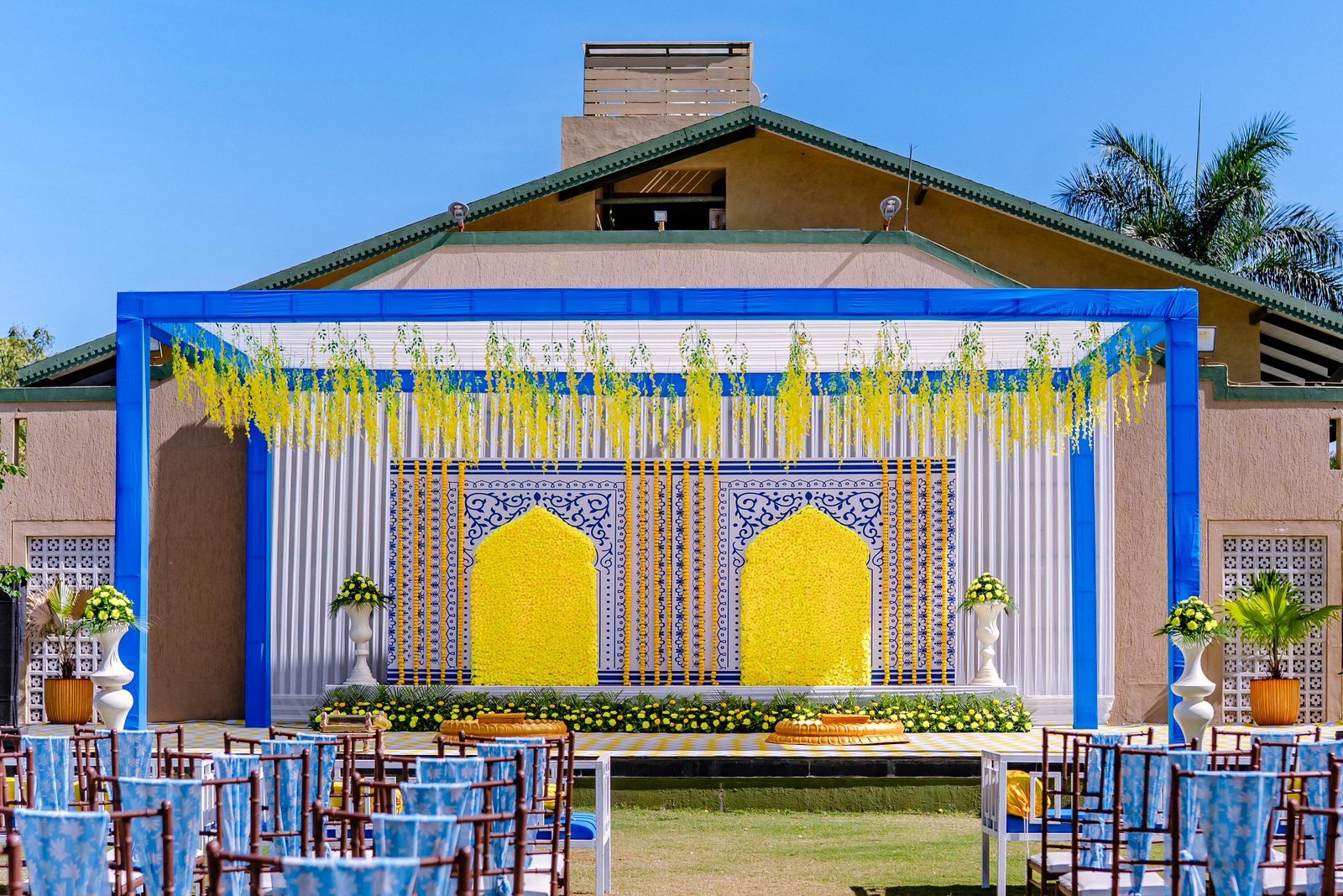 Harit-Lila lawn by day: a blue-and-yellow haldi stage on the lawn with the stage building behind